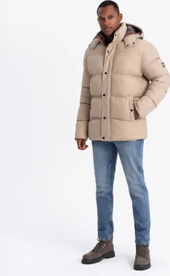 A man wearing a beige puffer jacket and blue jeans stands against a white background.