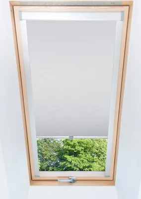 A skylight window with a light gray roller blind partially covering the view of green trees outside.