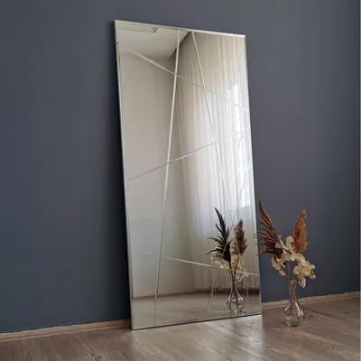A full-length mirror with a geometric design, leaning against a dark gray wall. Two vases with dried flowers are placed on the floor in front of the mirror.