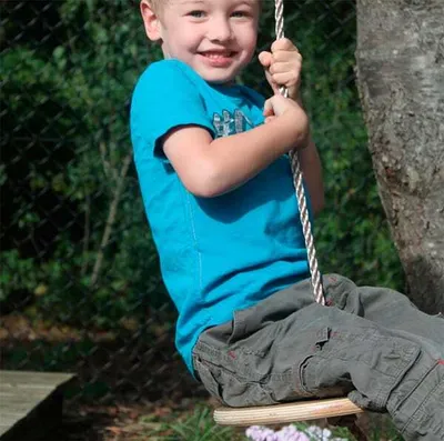 A young boy is sitting on a wooden swing, holding onto the rope, and smiling.