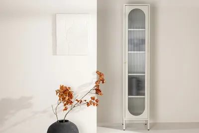 A tall, narrow beige display cabinet with glass doors and shelves, placed against a white wall. A dried floral arrangement in a dark vase is in the foreground.