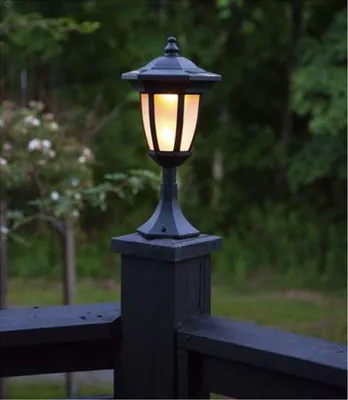 A black outdoor lamp post with a lit flame-shaped bulb, mounted on a wooden deck railing.