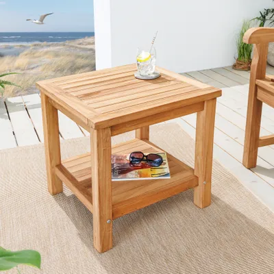A wooden side table with a drink and sunglasses on it, placed on a rug outdoors with a beach scene in the background.