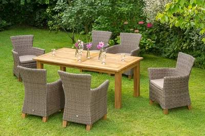 A dining set with a wooden table and wicker chairs is arranged on a grassy lawn in a garden setting, surrounded by lush greenery and flowers.