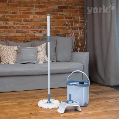 A mop and bucket set are placed on a wooden floor in front of a sofa and a brick wall.