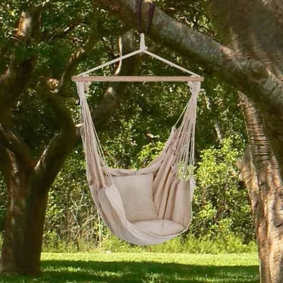 A beige hammock chair with a pillow hangs from a tree branch in a grassy outdoor setting.