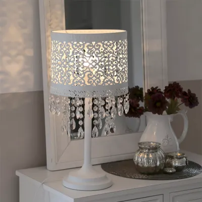 A white table lamp with an oriental pattern and crystal pendants sits on a white dresser next to a white pitcher with flowers and two decorative jars.
