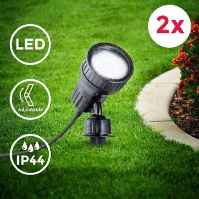 A close-up shot of a black LED spotlight placed in the grass, with decorative landscaping in the background. There are several informational icons and text overlays on the image.