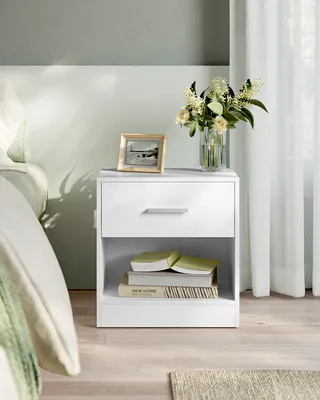 A white nightstand with a drawer and an open shelf, placed next to a bed. It holds a framed picture and a vase of flowers on top. Books are stacked in the open shelf.