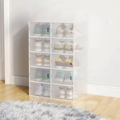 A stack of transparent shoe storage boxes filled with shoes, placed on a wooden floor next to a wall.