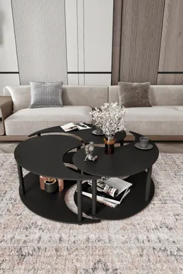A black, Yin Yang-shaped coffee table is shown in a modern living room setting with a light-colored sofa and decorative panels on the wall. The table has two tiers and holds a vase of flowers, books, and cups.