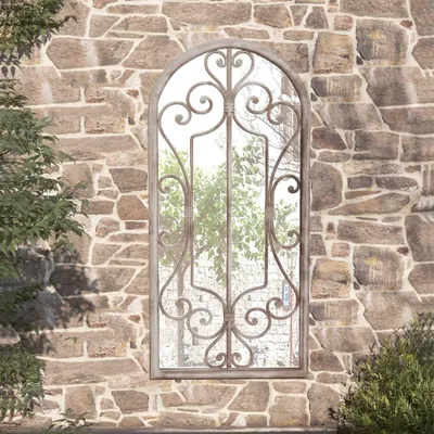 A decorative mirror with an ornate metal frame is mounted on a rustic stone wall, with greenery visible in the reflection and to the sides.