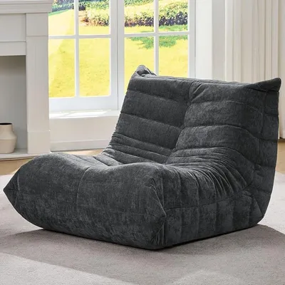 A dark gray bean bag chair is placed on a light gray carpet in front of a large window with a view of greenery.