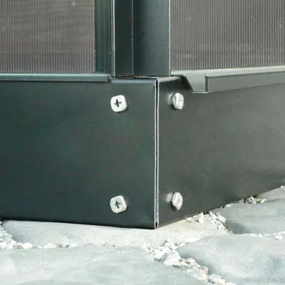 A close-up view of the corner of a dark gray metal structure, possibly a base or frame, with visible screws securing two panels together. The structure sits on a textured gray surface, possibly paving stones or concrete, with small white gravel scattered a