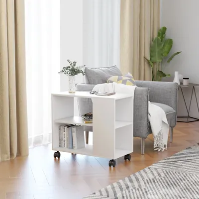 A white side table with shelves is placed next to a gray armchair in a living room setting. The table has books and a phone on it. Curtains are visible in the background.