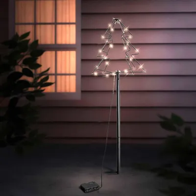A lit-up Christmas tree decoration in a garden at night, with a window and house siding in the background.