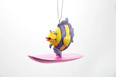 A cartoon fish ornament is shown on a pink surfboard, suspended by a chain against a white background.