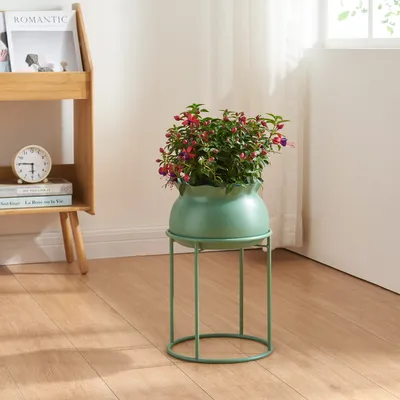 A green plant stand with a flowering plant in a green pot, placed on a wooden floor next to a bookshelf.