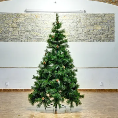 A green artificial Christmas tree with pinecones on a stand, placed indoors against a stone wall.