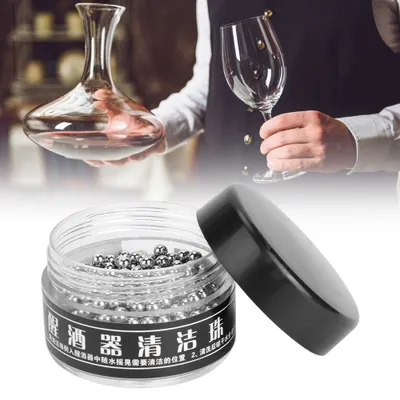 A person is holding a wine decanter and a wine glass, with a jar of cleaning balls in the foreground.
