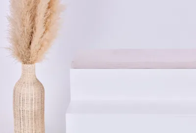 A close-up of a woven vase with dried pampas grass and a white tiered shelf.