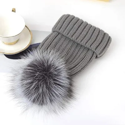 Graue Strickmütze mit großem schwarz-weißem Kunstfell-Pompon, neben einer weißen Teetasse