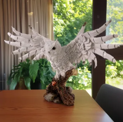 A white owl sculpture with outstretched wings is perched on a branch with rocks, placed on a wooden table in front of a window with greenery visible outside.