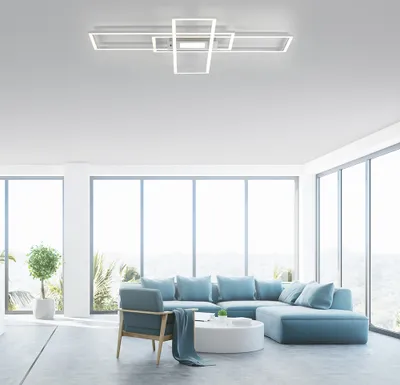 A modern living room with a sectional sofa, armchair, and coffee table, illuminated by a contemporary ceiling light fixture. Large windows offer a view of a scenic landscape.