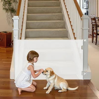A young girl and a puppy interact at the bottom of a staircase, separated by a white mesh baby gate.
