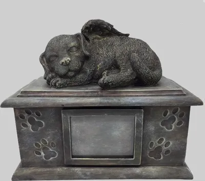 A pet urn shaped like a sleeping dog with wings, resting on a box with paw prints and a picture frame.