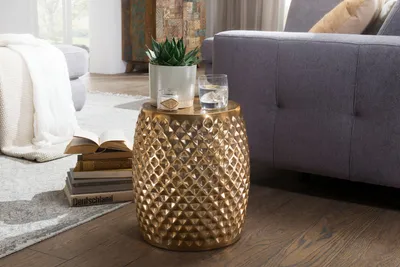 A gold drum-shaped side table with a textured surface is placed next to a gray sofa. A potted plant and a glass of water sit on top of the table. A stack of books is on the floor next to the table.