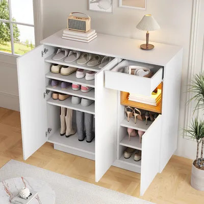 A white shoe cabinet with a drawer and open doors, displaying various shoes and boots, placed in a room with a window and a plant.