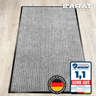 A gray doormat with vertical stripes is shown on a wooden floor. There are marketing overlays and logos on the image.