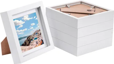 A white 3D box picture frame with a mat and a picture of a coastal town is shown on the left, and a stack of five identical frames is shown on the right.