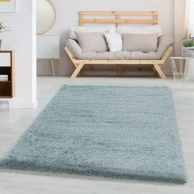 A light blue shaggy rug is placed on a light wooden floor in a modern living room. A wooden sofa with light-colored cushions is visible in the background.