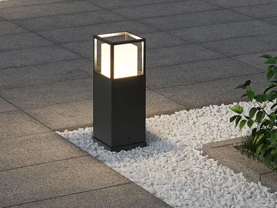 A modern black outdoor LED bollard light with a frosted glass diffuser, set in a garden with grey paving stones and white gravel.