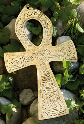 A golden ankh pendant with hieroglyphs is placed on a bed of rocks and green plants.