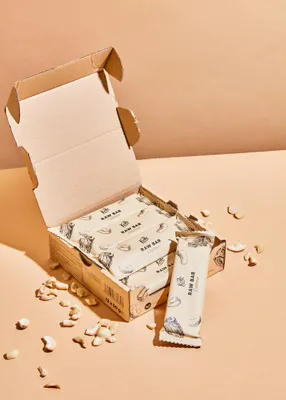 A cardboard box filled with individually wrapped raw cashew bars, with some loose cashews scattered around the box.