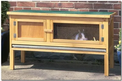 A wooden rabbit hutch with a green roof, placed on a patio in front of a brick wall.