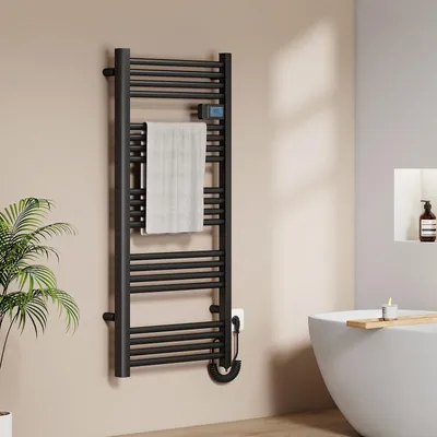 A black electric towel warmer is mounted on a light beige wall in a bathroom, with a white towel draped over it. A plant is visible to the left, and a white bathtub with a wooden tray and a candle is to the right.