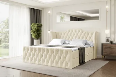 A bedroom scene with a large bed as the focal point, decorated with pillows and a throw blanket. A nightstand with decorative items is visible to the right, and a large window with sheer curtains is to the left.