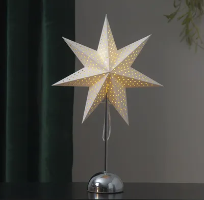A white star-shaped lamp with small perforations, lit from within, sits on a dark surface. Behind it, a dark green curtain is visible.