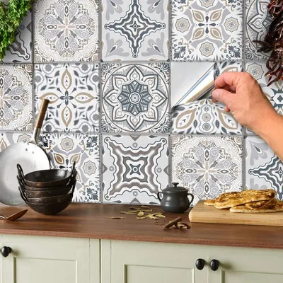 A hand is shown peeling a tile sticker from a wall covered in patterned tile stickers, with kitchen items on a countertop below.
