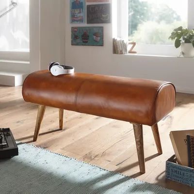 A brown leather bench with wooden legs in a room with a window and decorative items.
