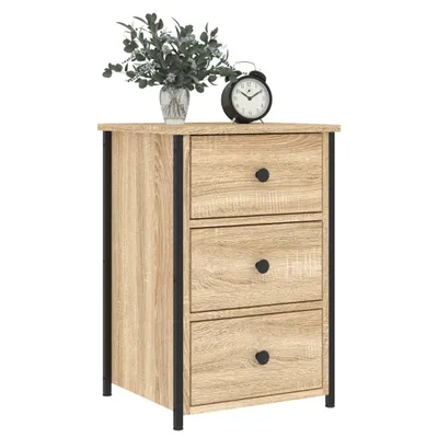 A light brown wooden nightstand with three drawers and black metal frame. It has a bouquet of faux greenery and a black alarm clock on top.