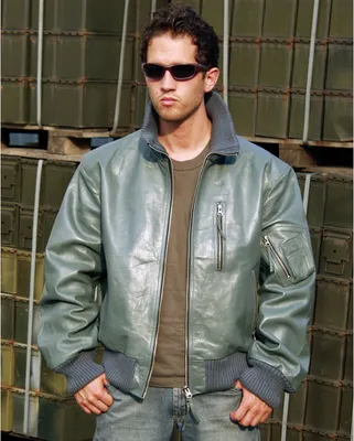 A man wearing a gray leather bomber jacket and sunglasses stands in front of stacked green crates.
