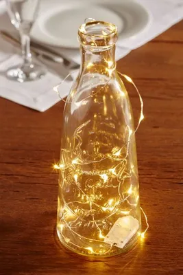 A clear glass bottle filled with white LED string lights sits on a wooden table.