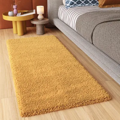 A yellow rug is placed on a wooden floor next to a bed and a small side table with decorative items.