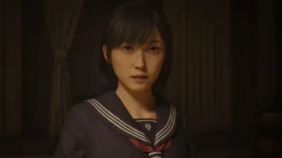 A young woman in a school uniform looks directly at the camera in a dimly lit room.