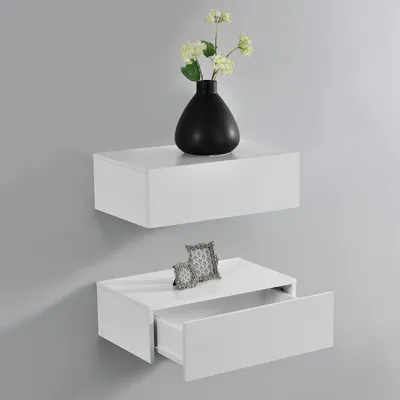 Two white floating shelves mounted on a gray wall. The top shelf holds a black vase with white flowers. The bottom shelf has a drawer partially open and a small decorative picture frame on top.